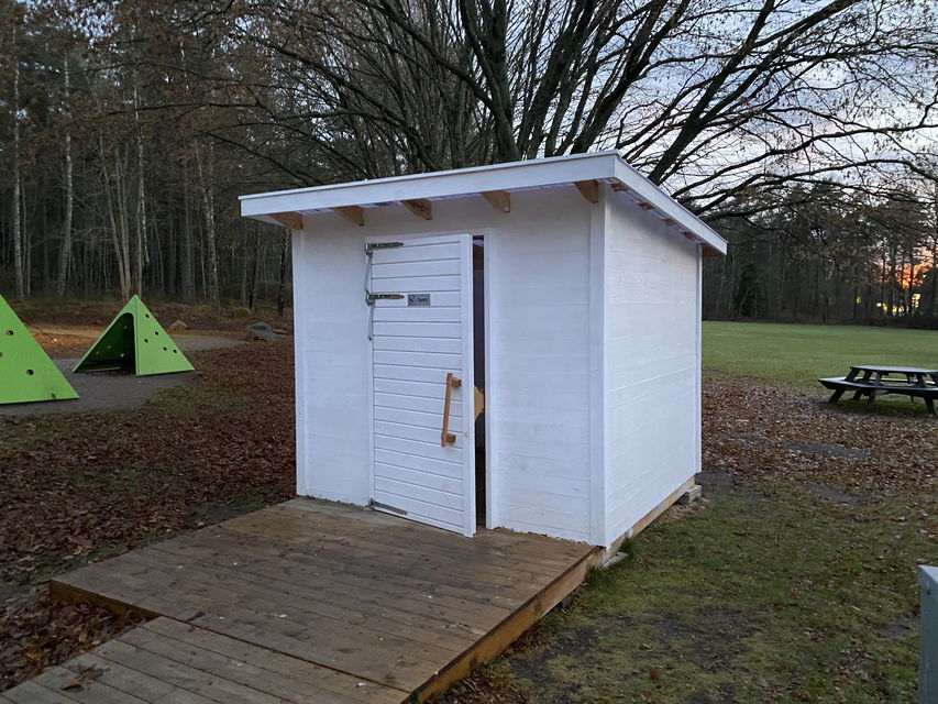 Vitmålat toaletthus med ramp framför. Höstbild.