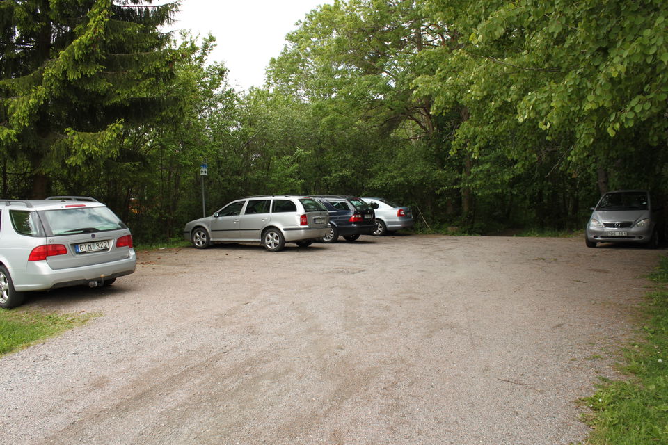 Fem bilar står på en grusad parkeringsplats med skog i bakgrunden. Det finns utrymme för flera bilar till på parkeringen.