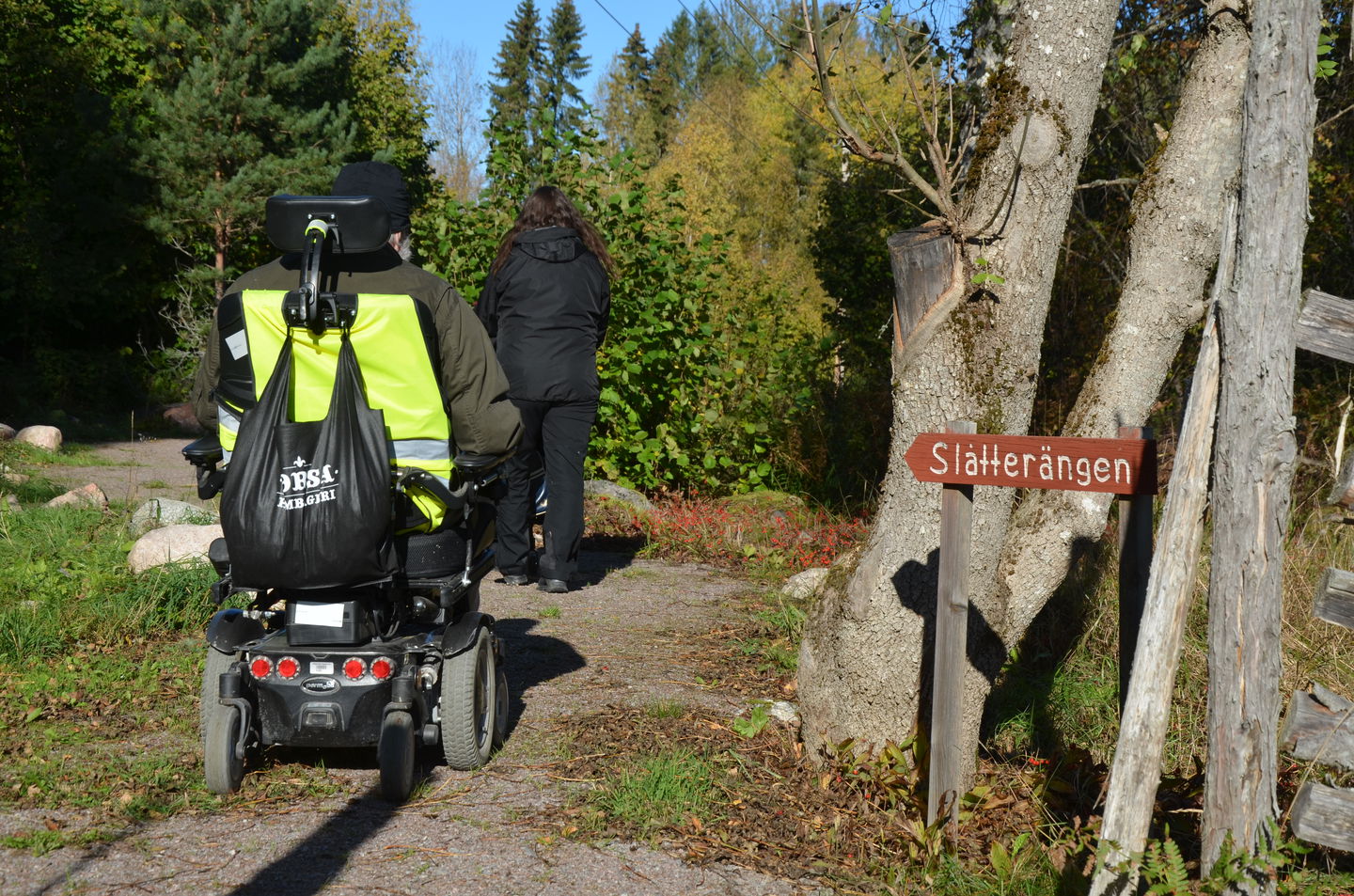 En person i elrullstol sedda bakifrån och en gånde framför, på den hårdgjorda stigen. Lummig grönska, lite höstigt.