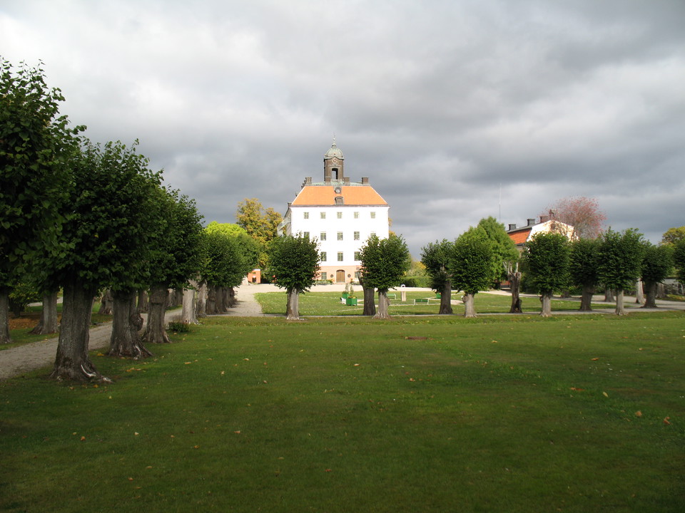 Ängsö slott med slottsträdgården framför. Himlen är grå men på det vita slottet skiner solen.