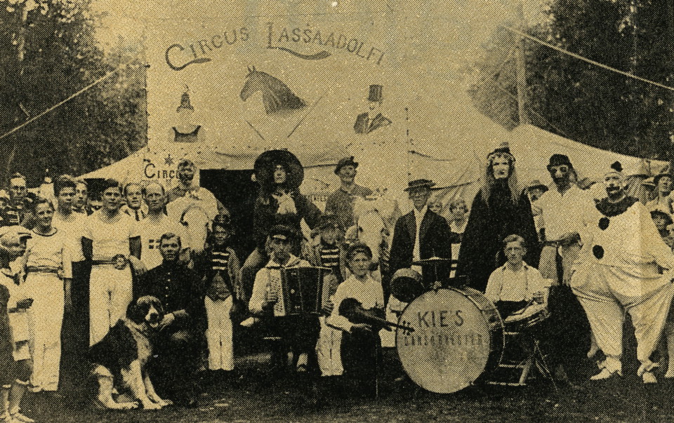 Cirkusartisterna på Cirkus Lassa Adolfi uppställda framför tältet. Några är klädda i gymnastikkläder och några i clownliknande kläder. På bilden syns även musiker med trumset och dragspel, och en hund. 