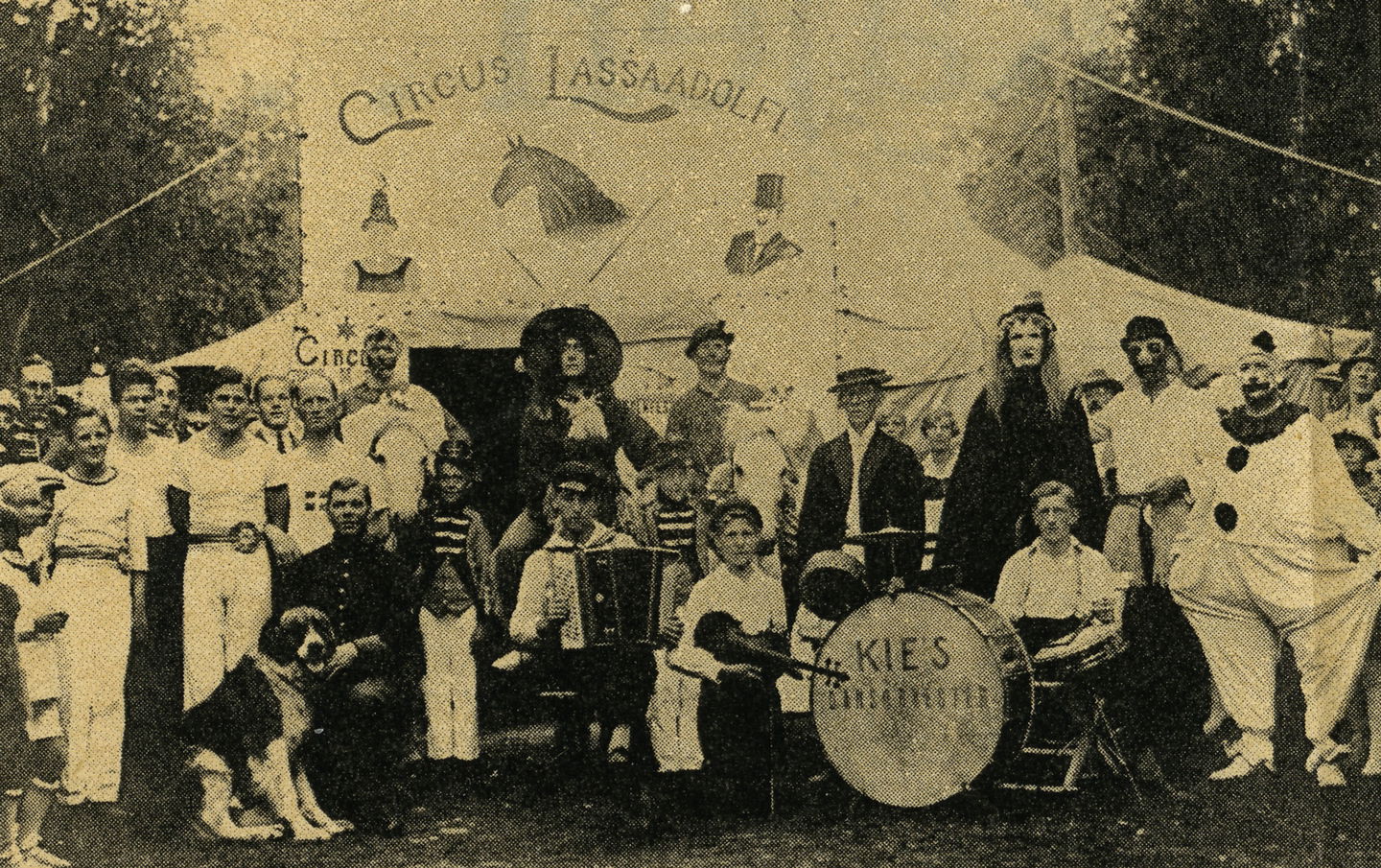 Cirkusartisterna på Cirkus Lassa Adolfi uppställda framför tältet. Några är klädda i gymnastikkläder och några i clownliknande kläder. På bilden syns även musiker med trumset och dragspel, och en hund. 