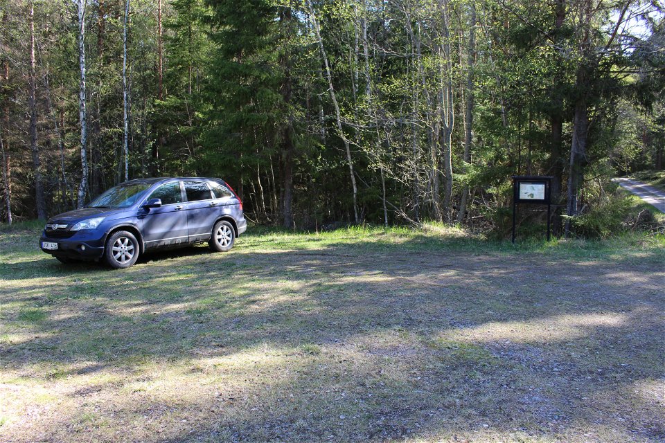 En bil står på en grusad parkeringsplats med skog bakom. På platsen finns en informationstavla och i bredvid går en skogsbilväg.