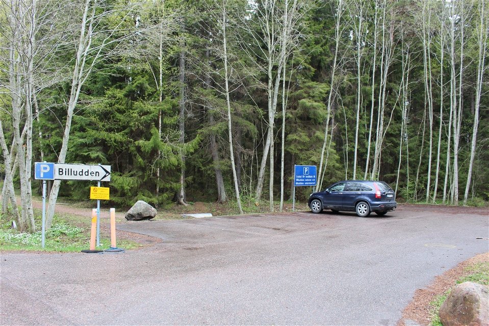 En bil står på en asfalterad parkering vid en bilväg. Vid parkeringen finns en skylt med texten Billudden som pekar åt höger.