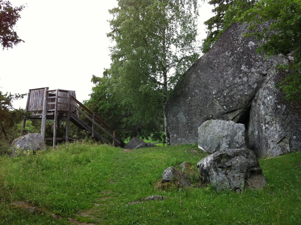 Ett litet fågeltorn står bredvid en gräsklädd stig och en stor stenbumling. En trappa leder upp till tornets plattform.