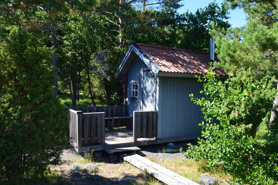En smal spång leder fram till en altan framför en liten bastu. Det är ett lågt trappsteg upp till altanen som har ett räcke runtom och en sittbänk.