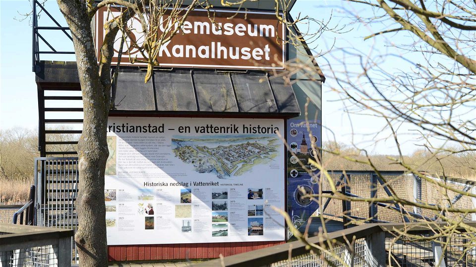 Kanalhuset framsida med en stor brunvit skylt med texten Utemuseum Kanalhuset. Under skylten täcks väggen av bild och text som berättar om staden Kristianstads vattenrika historia.