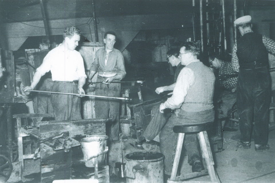 Män som arbetar i glashyttan på Elme glasbruk. Några snurrar blåsrör med glas på, en man sitter på en pall intill. 