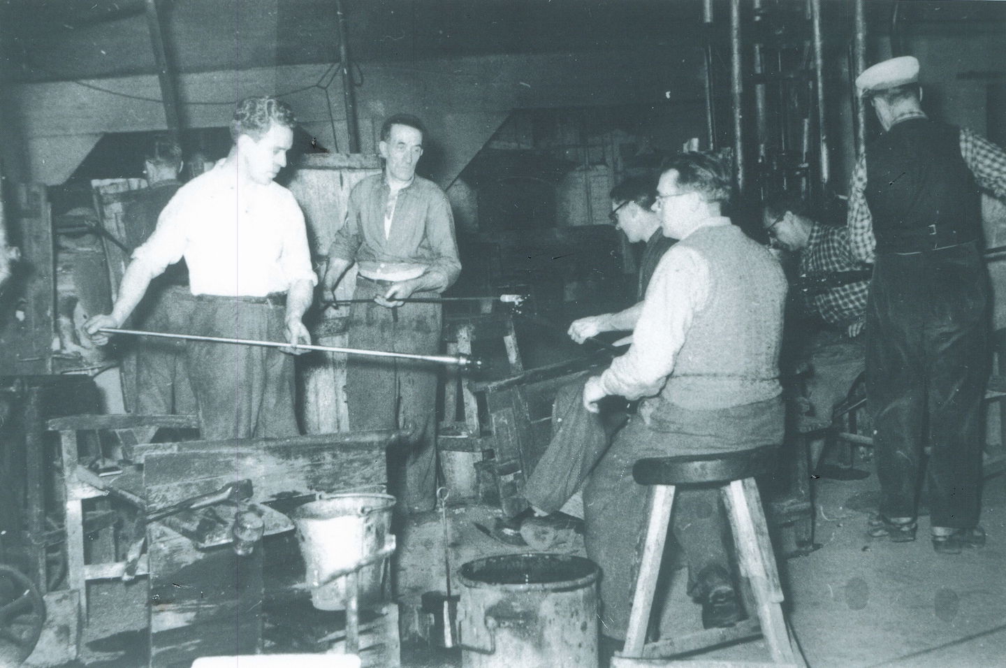 Män som arbetar i glashyttan på Elme glasbruk. Några snurrar blåsrör med glas på, en man sitter på en pall intill. 