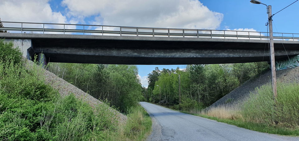En bilväg rakt fram. Bilvägen korsas av en viadukt. Natur kantar vägen.