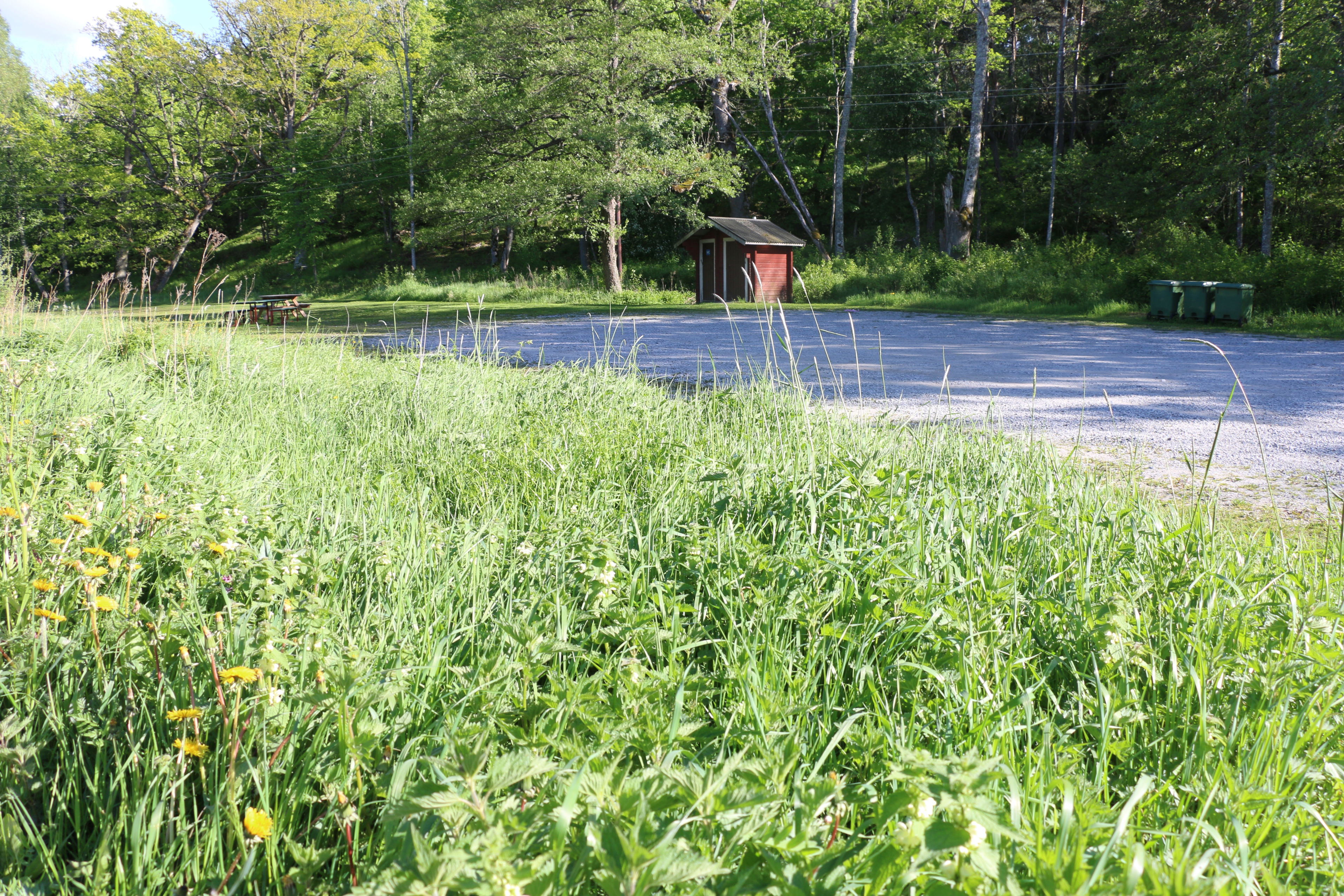 Grusad parkeringsplats i grönskande landskap. Liten röd byggnad bredvid parkeringen.
