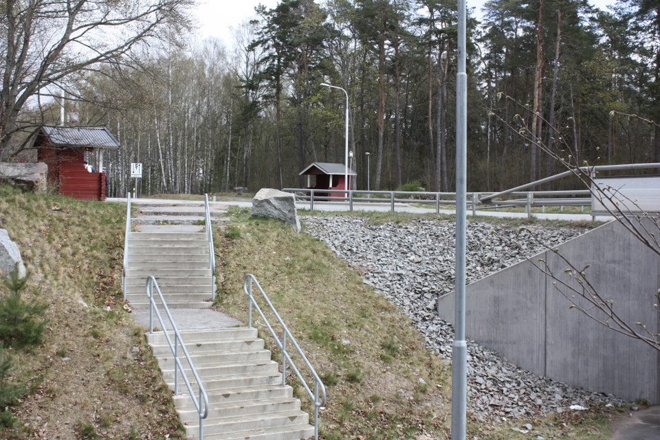 En stentrappa med handräcken leder upp till en bilväg. På båda sidorna av vägen står en busskur med skylt för busshållplats. Det finns gatubelysning vid trappan och busskurarna.
