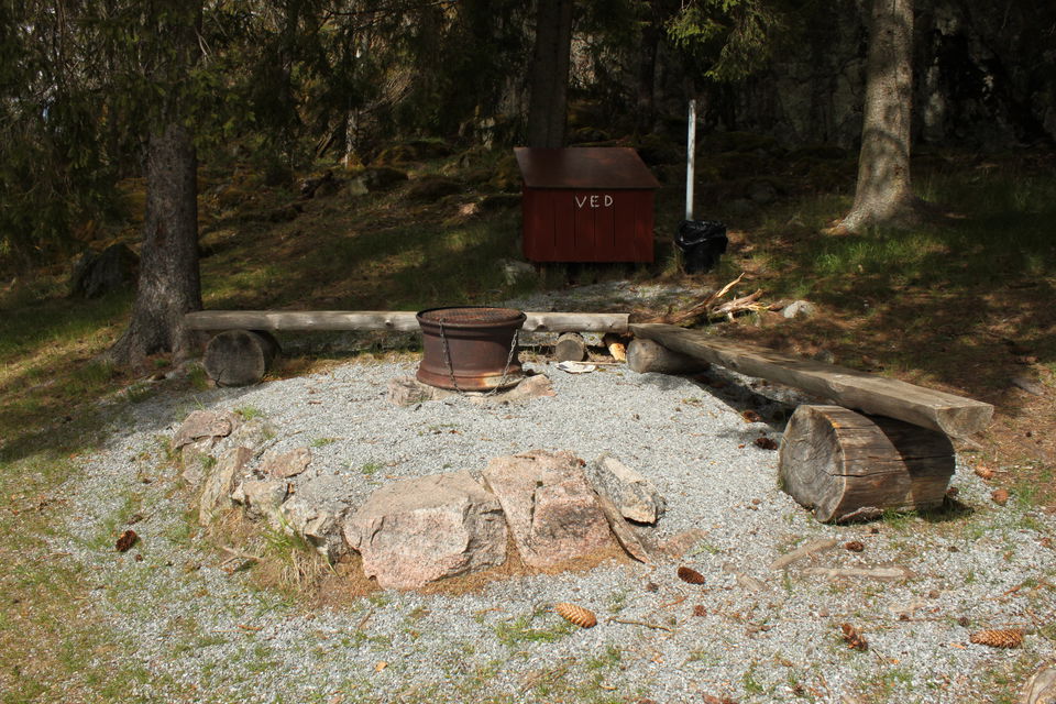 En grusad grillplats med sittbänkar och ett litet vedförråd en bit bort.