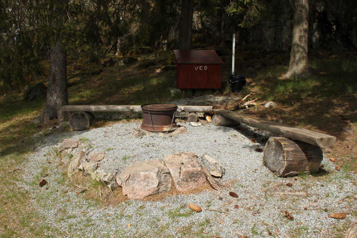 En grusad grillplats med sittbänkar och ett litet vedförråd en bit bort.