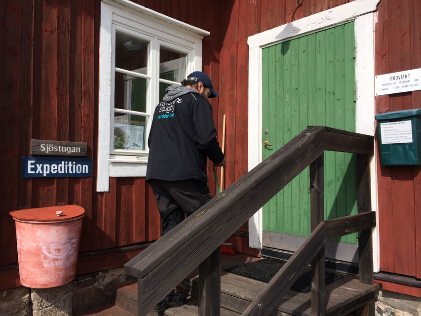 En man går uppför en trappa med handräcken upp till en entrédörr. På väggen finns skyltar med texten "Sjöstugan" och "Expedition".