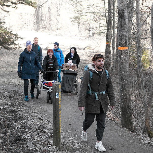 Sju personer promenerar på en bred stig i skogen. En går med rollator och en blir skjutsad i rullstol.