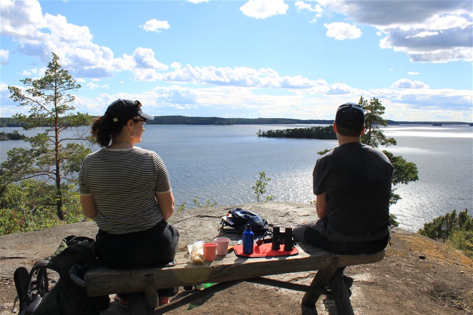 En man och en kvinna sitter på en bänk på ett högt berg. De har dukat fram fika. Från platsen har de utsikt över vattnet.