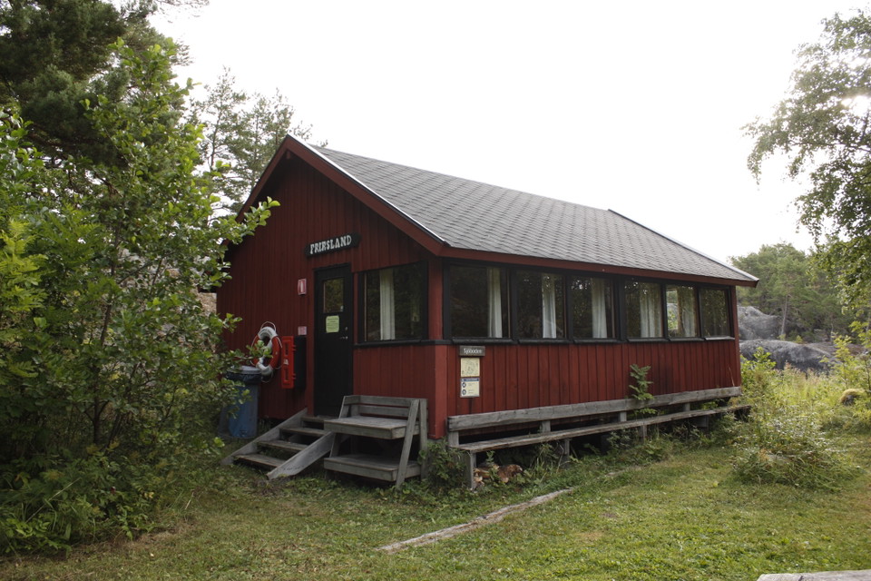En bod med sittbänkar utanför och en låg trappa upp till ingången. Runtom finns träd och bergshäll.