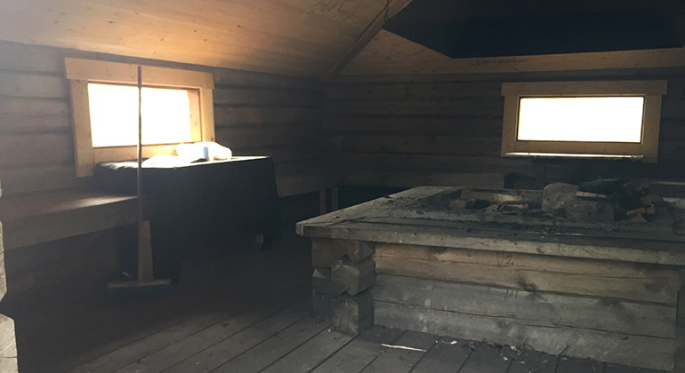 Inuti en raststuga med fönster bänkar och eldplats i mitten.