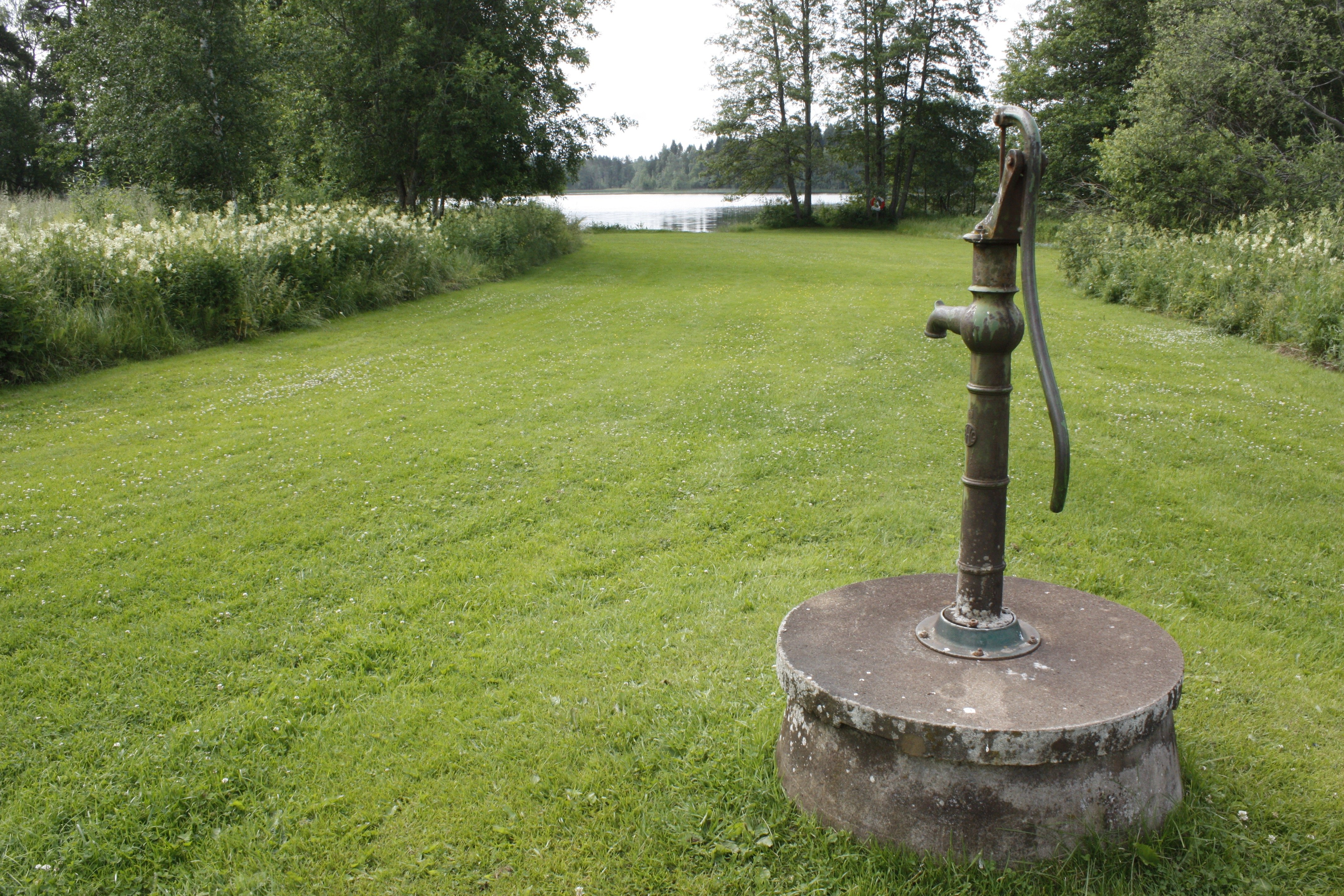 En gammaldags brunn med pump står på en välklippt gräsmatta. Gräsmattan leder till vattnet.
