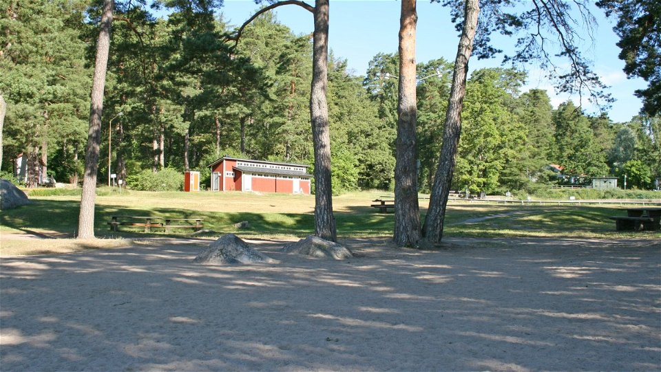 En sandstrand med träd och större stenar i utkanten. I bakgrunden står en toalettbyggnad och bakom den är det många träd.