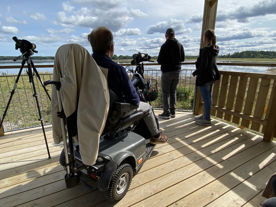 Tre personer varav en i rullstol  och en tubkikare. Utsikt över vattnet i bakgrunden.