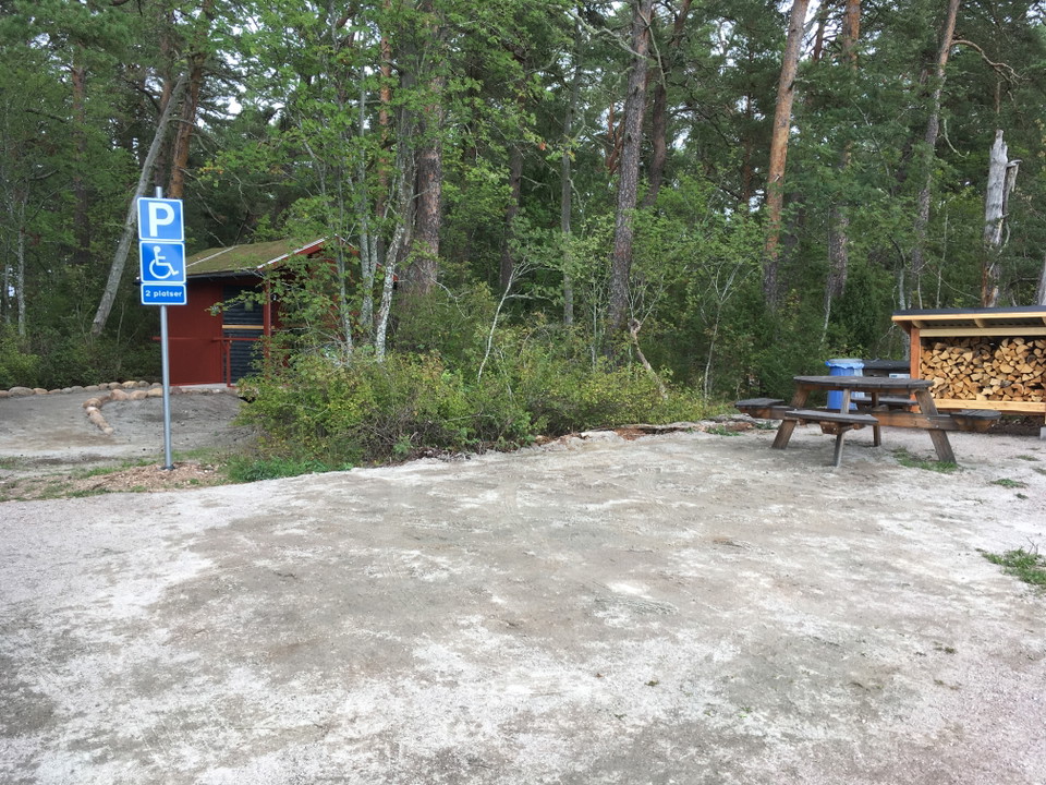 En jämn handikappparkering med torrtoalett, bänkbord och vedförråd i utkanten.