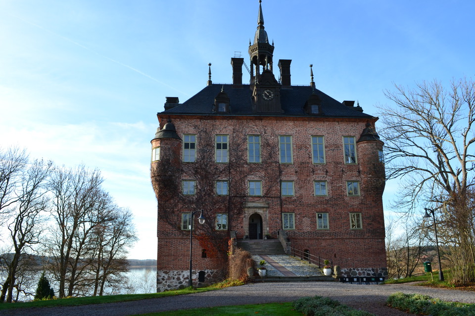 Ett slott med tre våningar och ett torn på taket. Framför slottet är finns en grusplan och en park. Bakom slottet syns vatten.