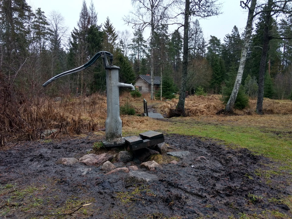 En vattenpump med dricksvatten står på lerig mark och är omringad av stenar. I bakgrunden syns något ojämn gräsmark och unsentorpet.