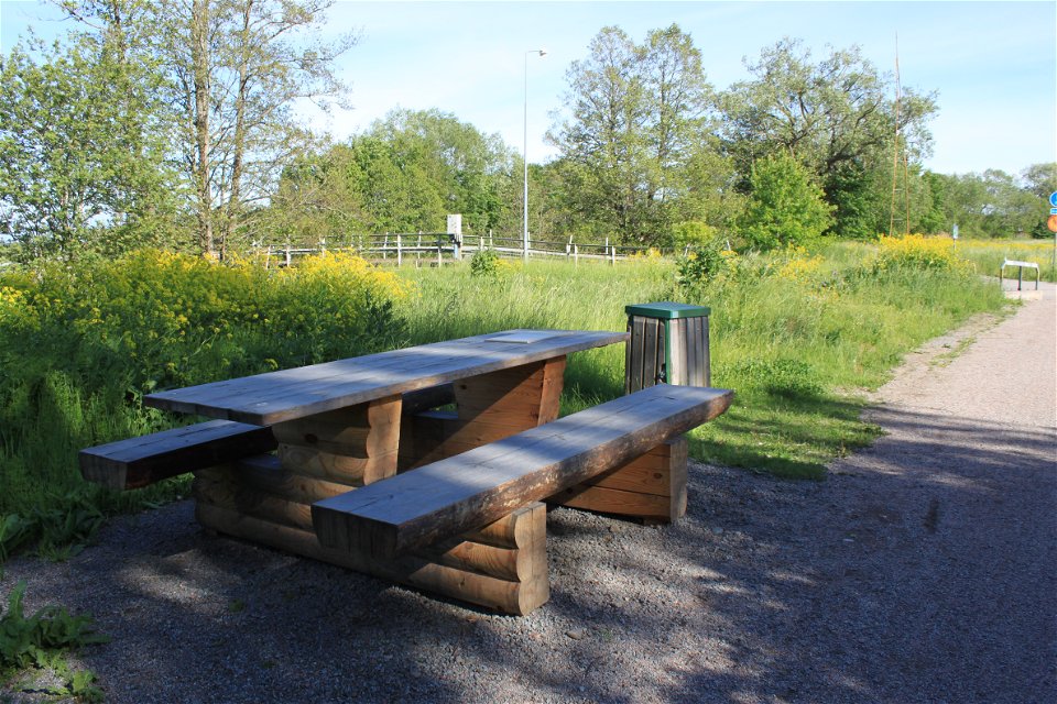 Ett bänkbord och en soptunna står vid sidan av en grusad gångväg. I bakgrunden finns en bro över Fyrisån.