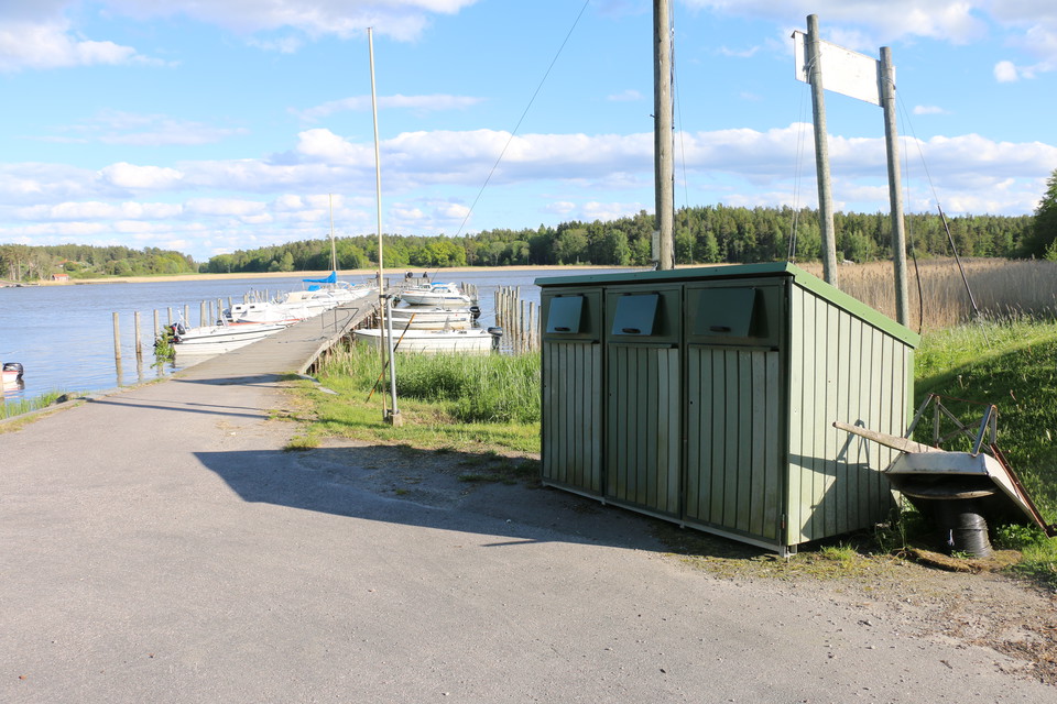 Båtbrygga med flera förtöjda båtar. På land finns flera soptunnor.
