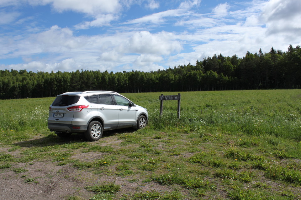 På en liten parkering med en parkeringsskylt står en bil. Underlaget är något ojämnt med grus och grästuvor