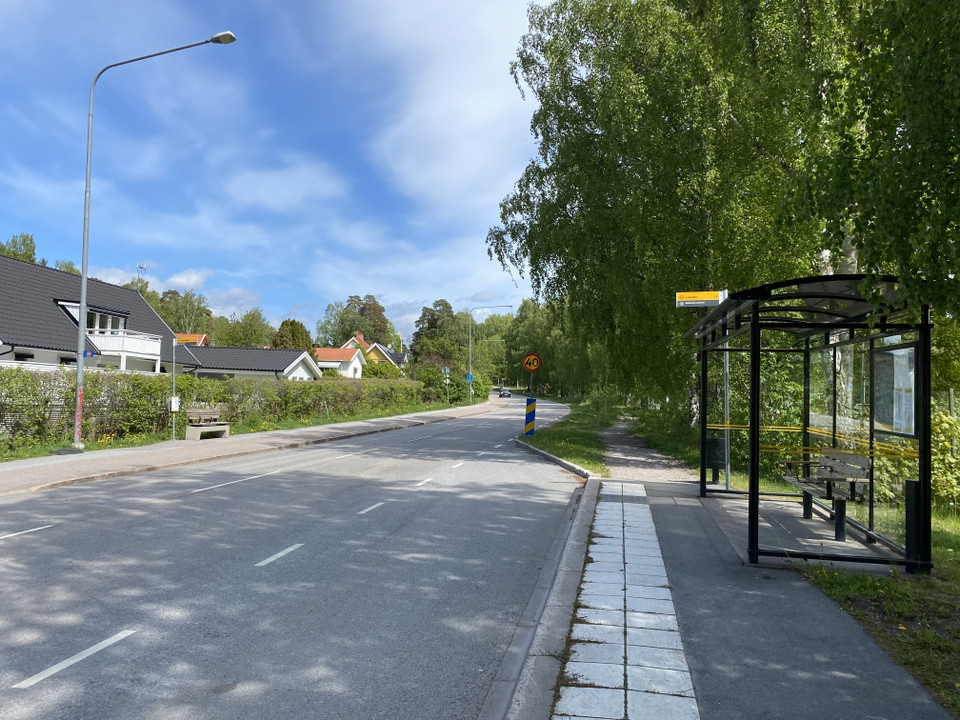 En busskur står på en busshållplats vid en bilväg. Mitt emot hållplatsen är det ett villaområde.