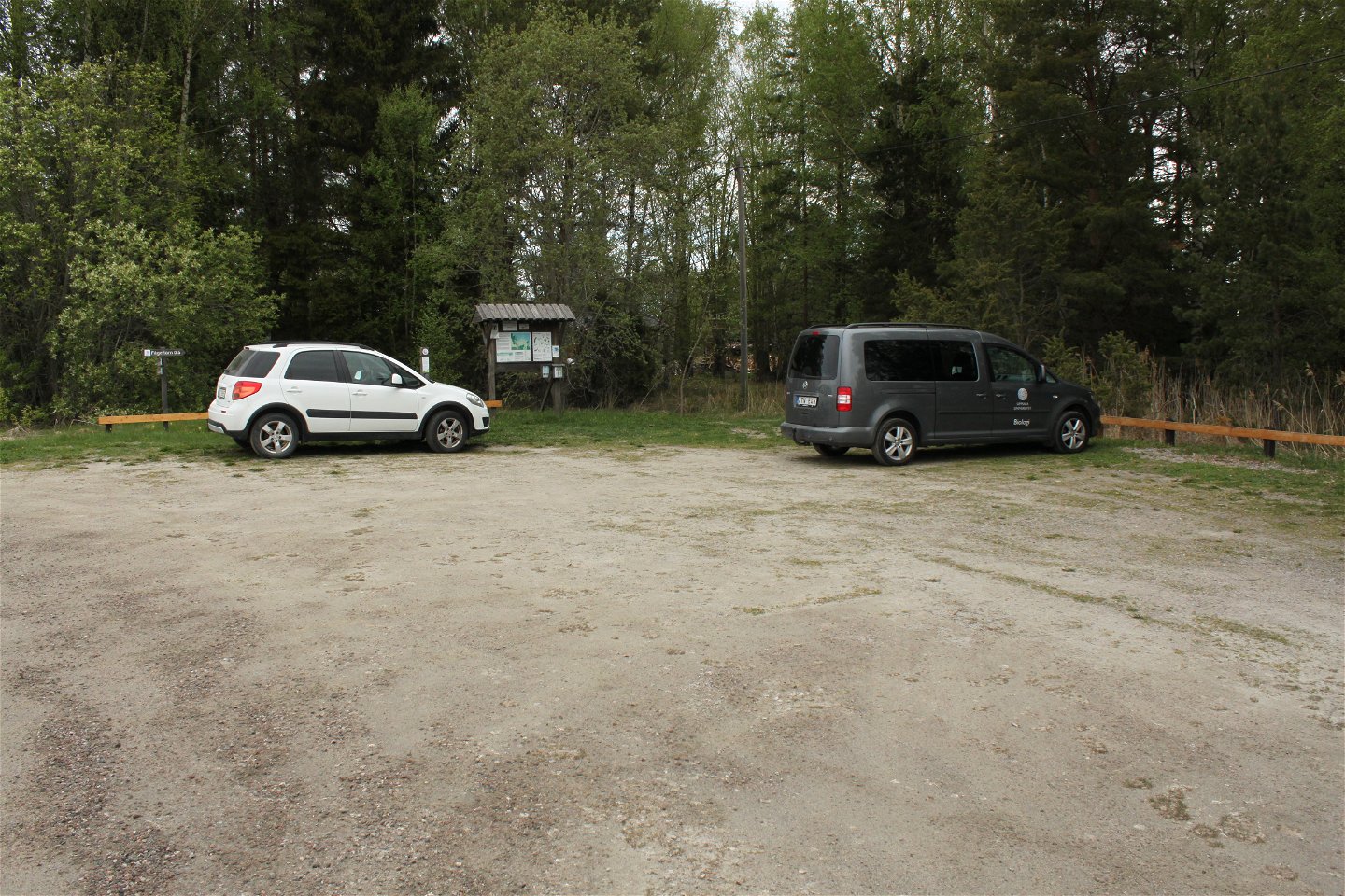 Två bilar står på en stor parkeringsplats. Bredvid parkeringen finns en infromationsskylt.