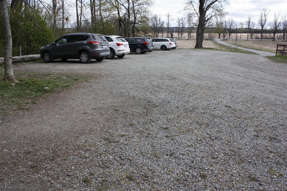 Sex bilar står på en grusad parkeringsplats. I bakgrunden syns en större bilväg och en grusväg som leder fram till parkeringen.