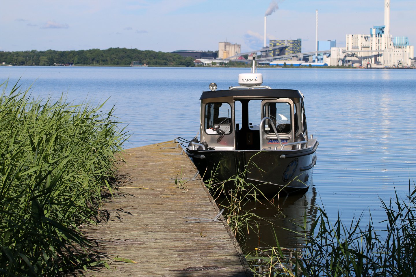 En båt ligger förtöjd vid en träbrygga. Västerås hamn skymtas i bakgrunden.