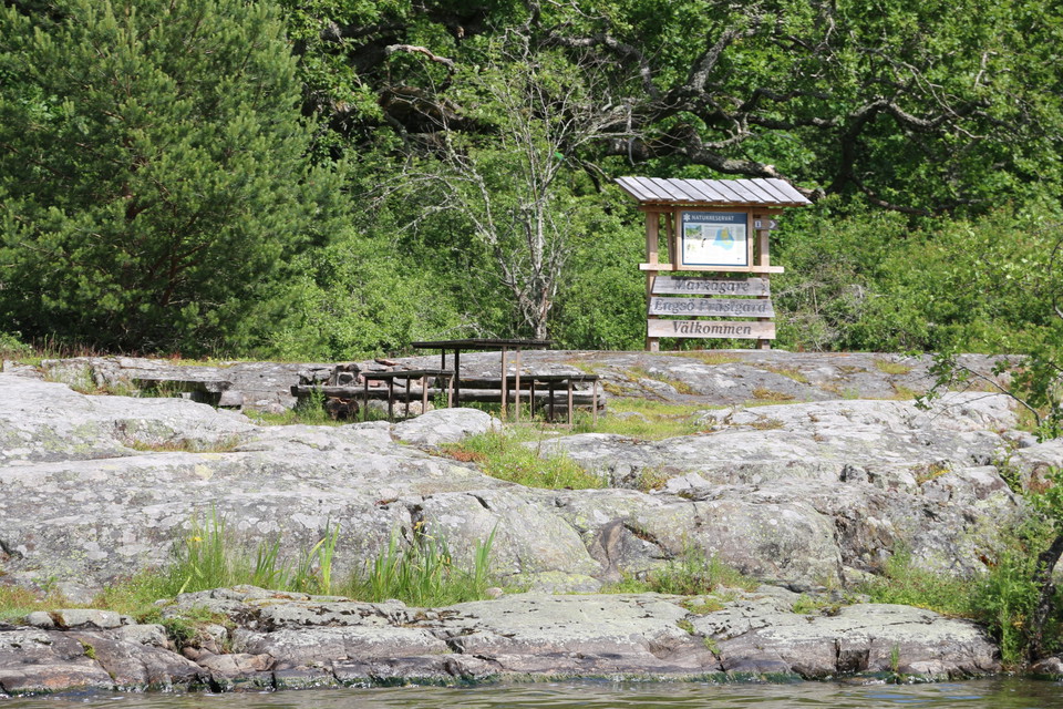 Rastplats med bänkar och bord på klippor intill Mälaren. Informationstavla i bakgrunden.