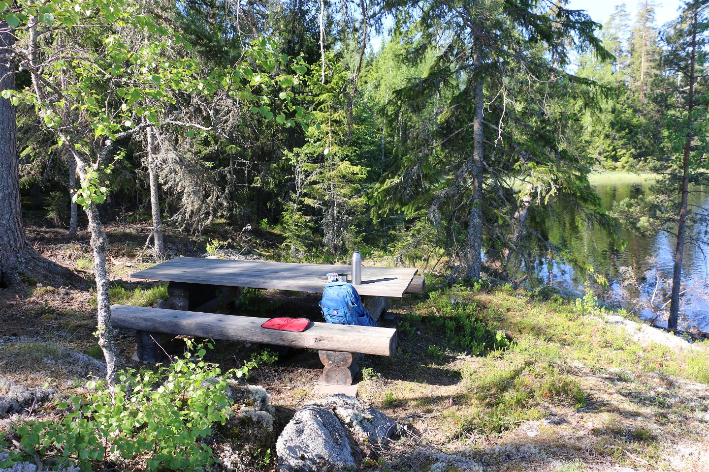 Ett picknickbord med en ryggsäck, ett sittunderlag, en termos, i skogen. En sjö skymtar i bakgrunden.