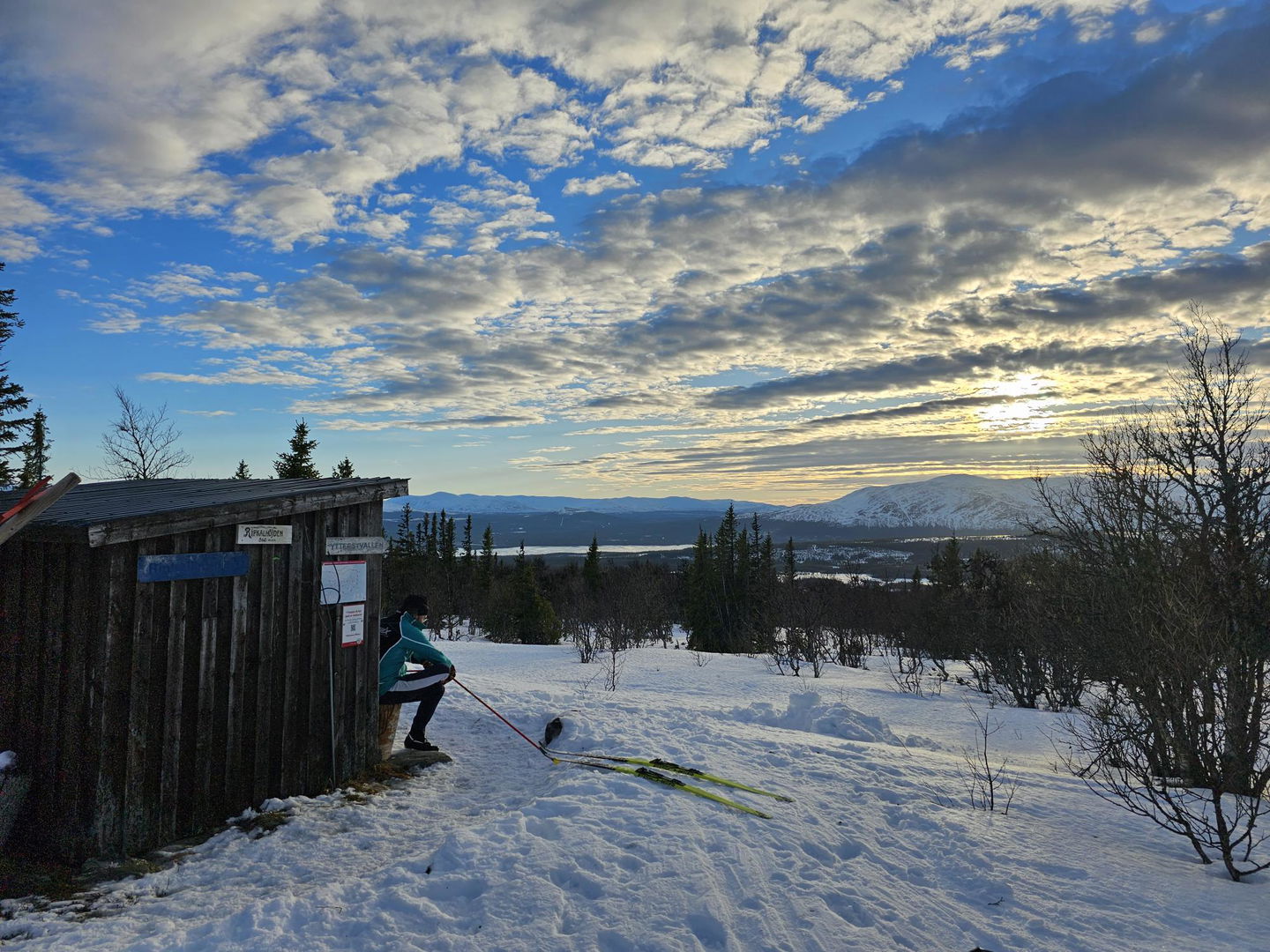 Person som sitter i ett vindskydd med skidor bredvid sig och milsvid utsikt framför sig