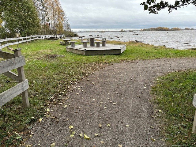 Grusgång som leder till en stand. Stranden är täckt av gräs. Det finns flera rastbänkar.