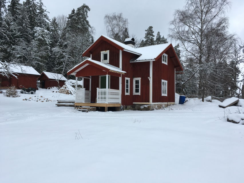 Ett torp står på en snötäckt öppen yta. Vid ena sidan av torpet står två mindre bodar.