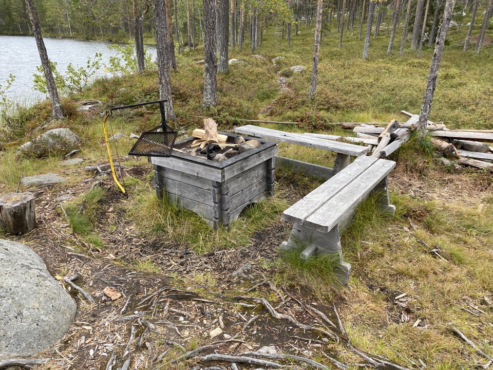 En eldstad med två bänkar i en skog vid en sjö. 