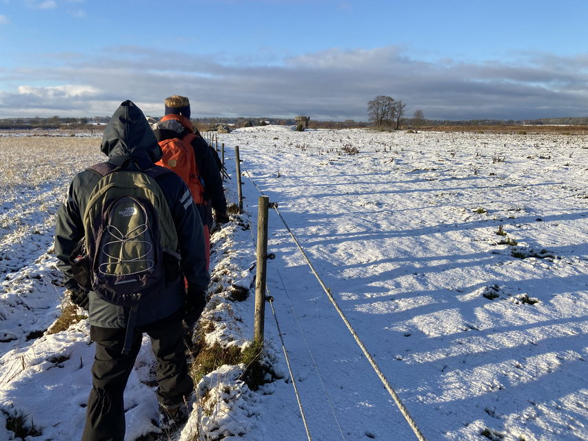 Två personer går på snötäckt mark vid sidan av ett staket. 