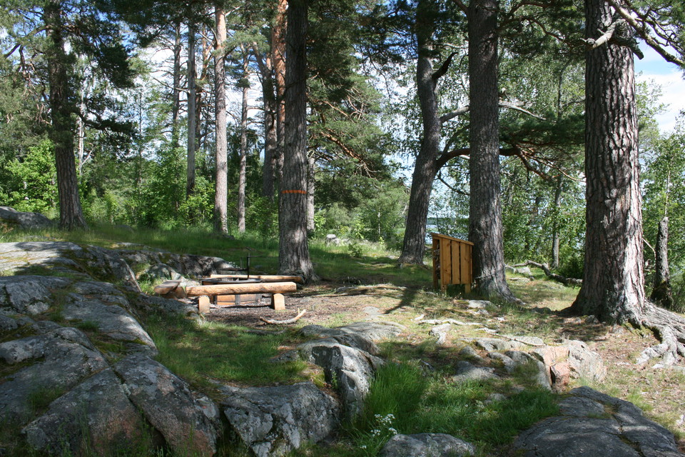 En grillplats med tre stockbänkar och en eldstad står bland klippor. Ett vedförråd still intill grillplatsen.