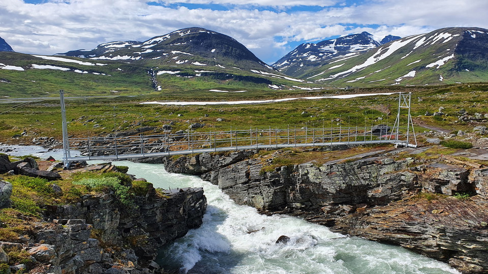 En bro i järn över en fjälljokk, med fjälltoppar i bakgrunden. 