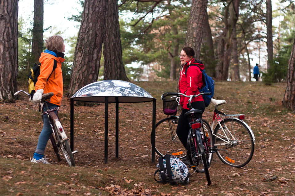 Två vuxna står vid en metallskulptur i skogen med tre cyklar. I bakgrunden går en person genom skogen.