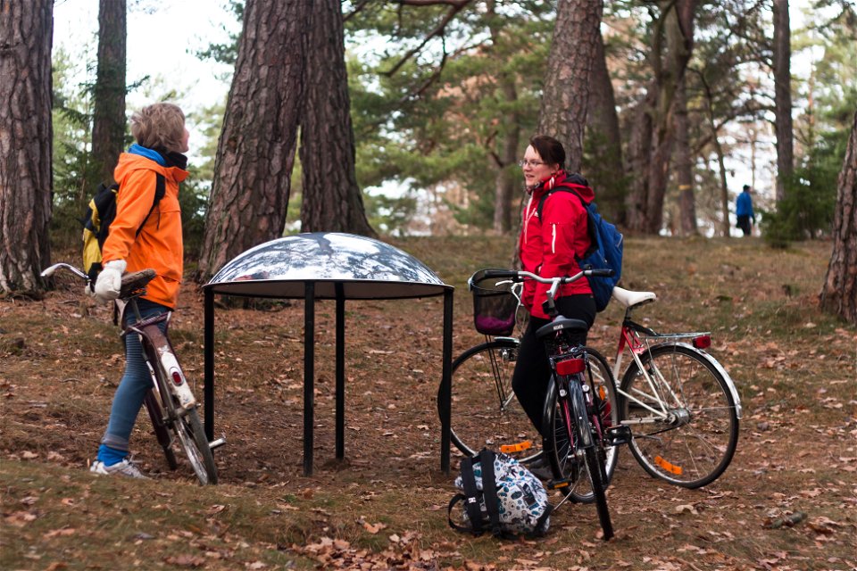 Två vuxna står vid en metallskulptur i skogen med tre cyklar. I bakgrunden går en person genom skogen.
