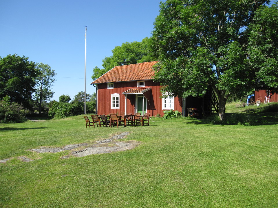 En stuga står på en gräsplan. Vid stugan står en flaggstång och framför står flera stolar och bord.
