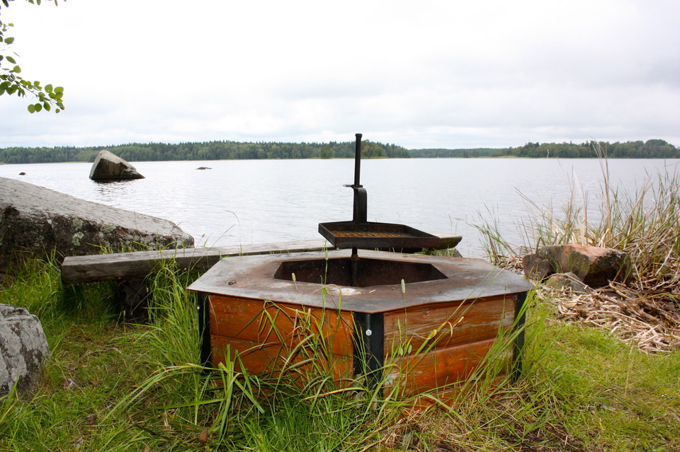 En grillplats med vridbart grillgaller och en låg sittbänk bredvid. Grillplatsen står vid en sjö.