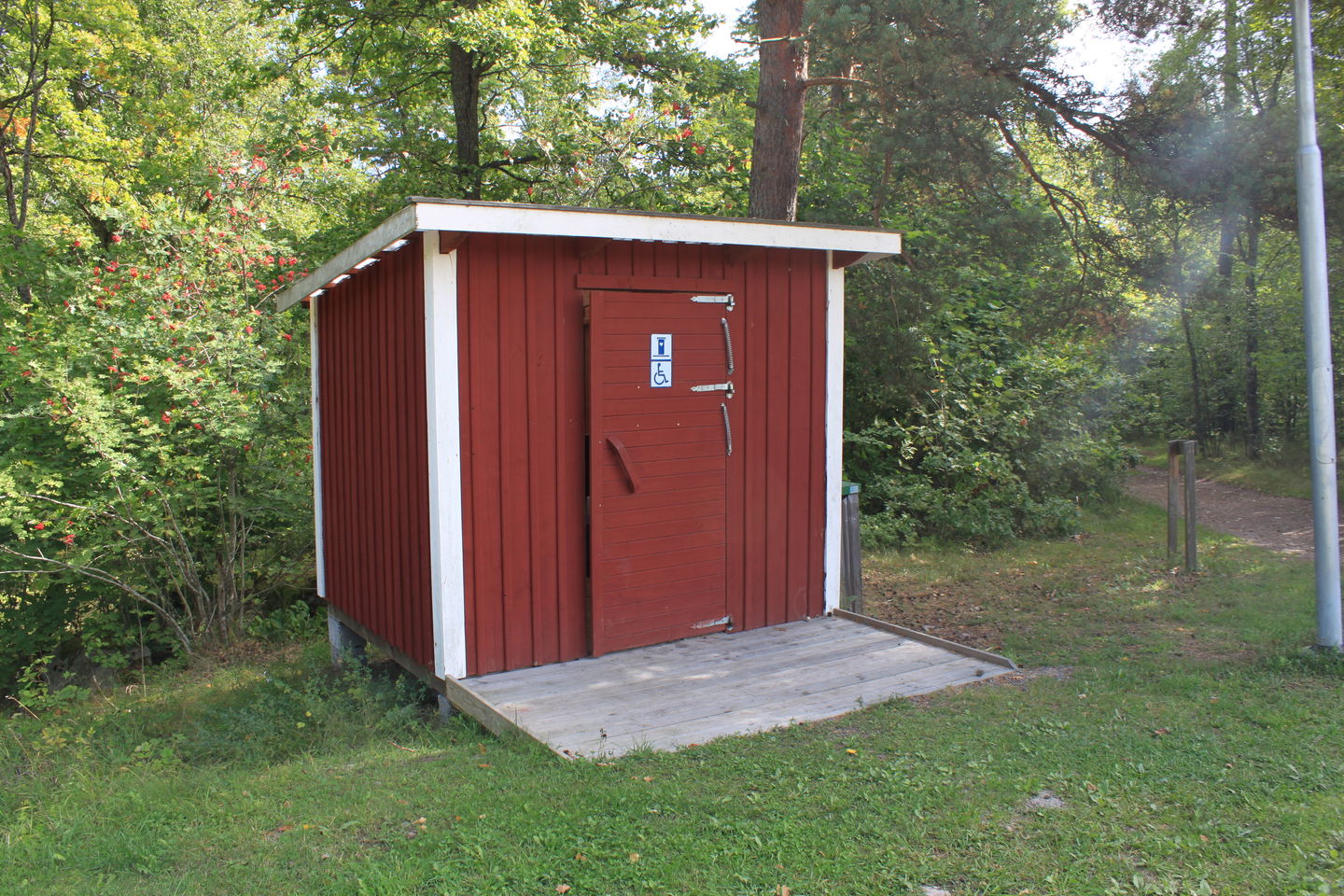 En tillgänglighetsanpassad torrtoalett står på en gräsmark. En kort ramp med kanter leder upp till toaletten.